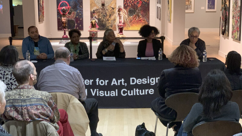 Photograph of a panel discussion in a gallery space. Five individuals are seated at a table, which is covered in a black table cloth. An audience is seated before them. Behind the panel are mixed media artworks, both hanging and on pedestals.