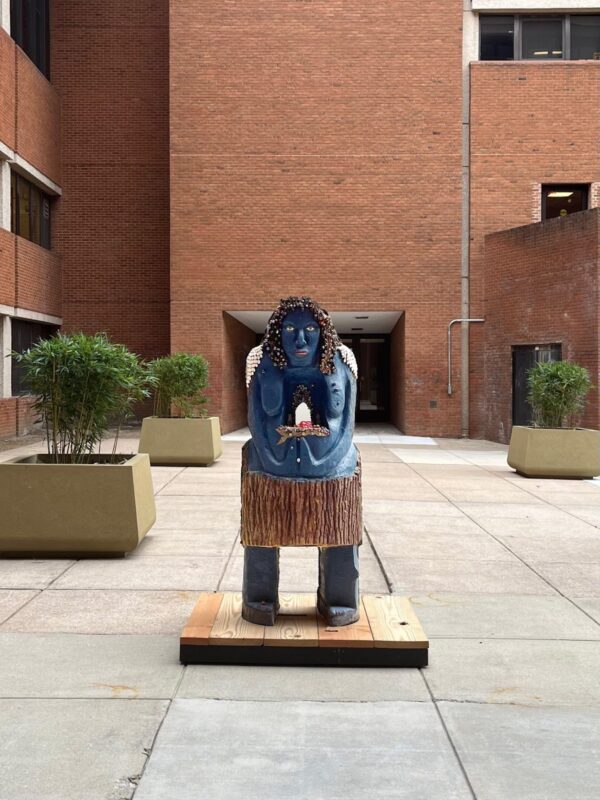 An abstract sculpture of a woman in blue. The subject is crafted from wood with a visible trunk around her waist. The work is placed upon a raw wooden pedestal in an outdoor space.