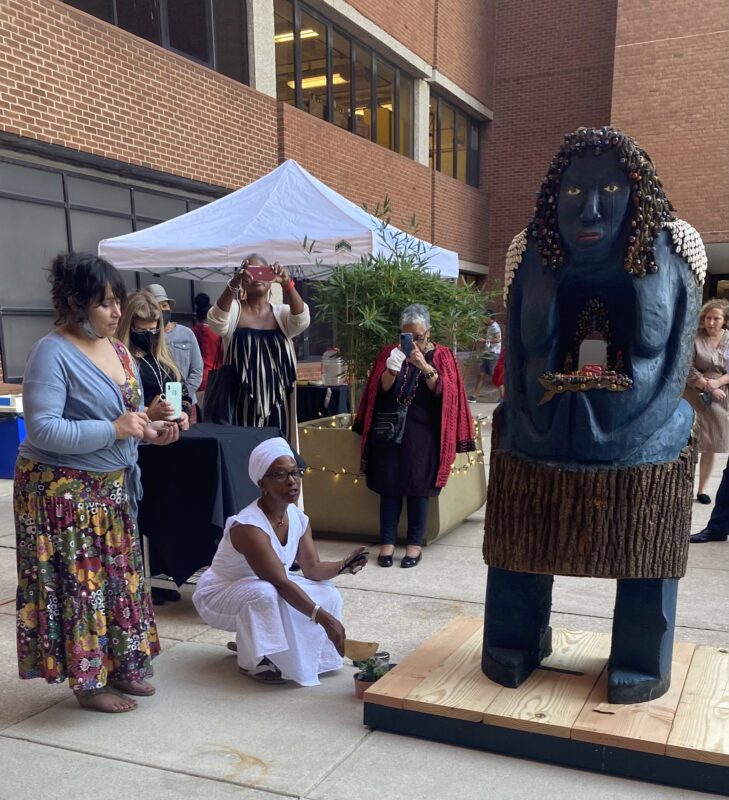 Several individuals gather around a blue wooden sculpture of a woman. A woman with a dark skin-tone dressed in white is crouched at the base of the sculpture. She holds a small piece of parchment in her white hand and waves it over burning incense.