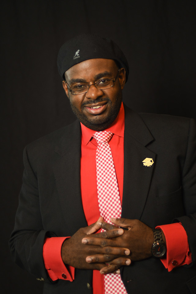 A portrait of a man with a dark skin-tone smiling for the camera. He wears a black beret, a black suit jacket, a red button-down shirt, and a red patterned tie. He clasps his hands together in front of his chest.