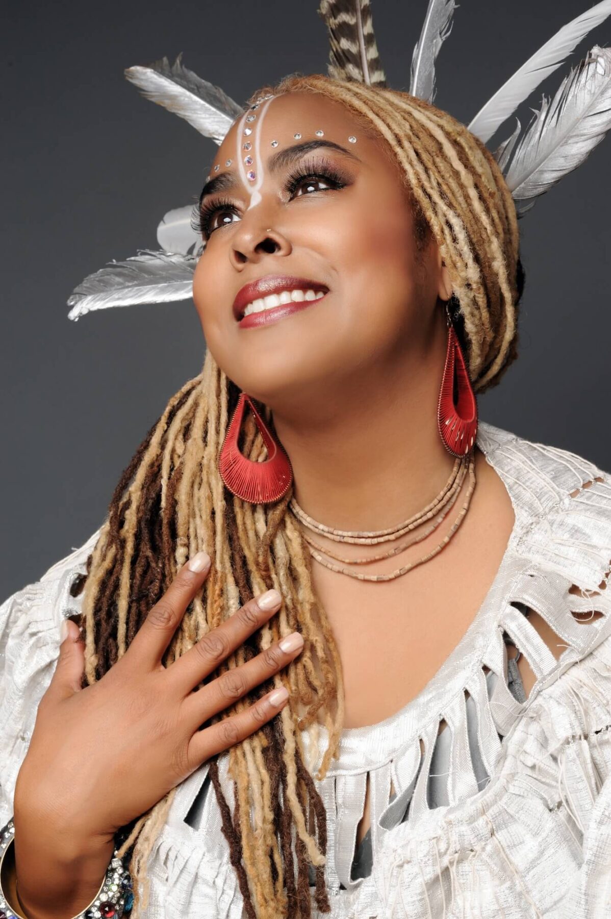 A portrait with a woman with a medium skin-tone with blonde and brown dreads. She wears white traditional face paint and white feathers in her hair. She is wearing a white fringed top and holds up her hand to her chest.