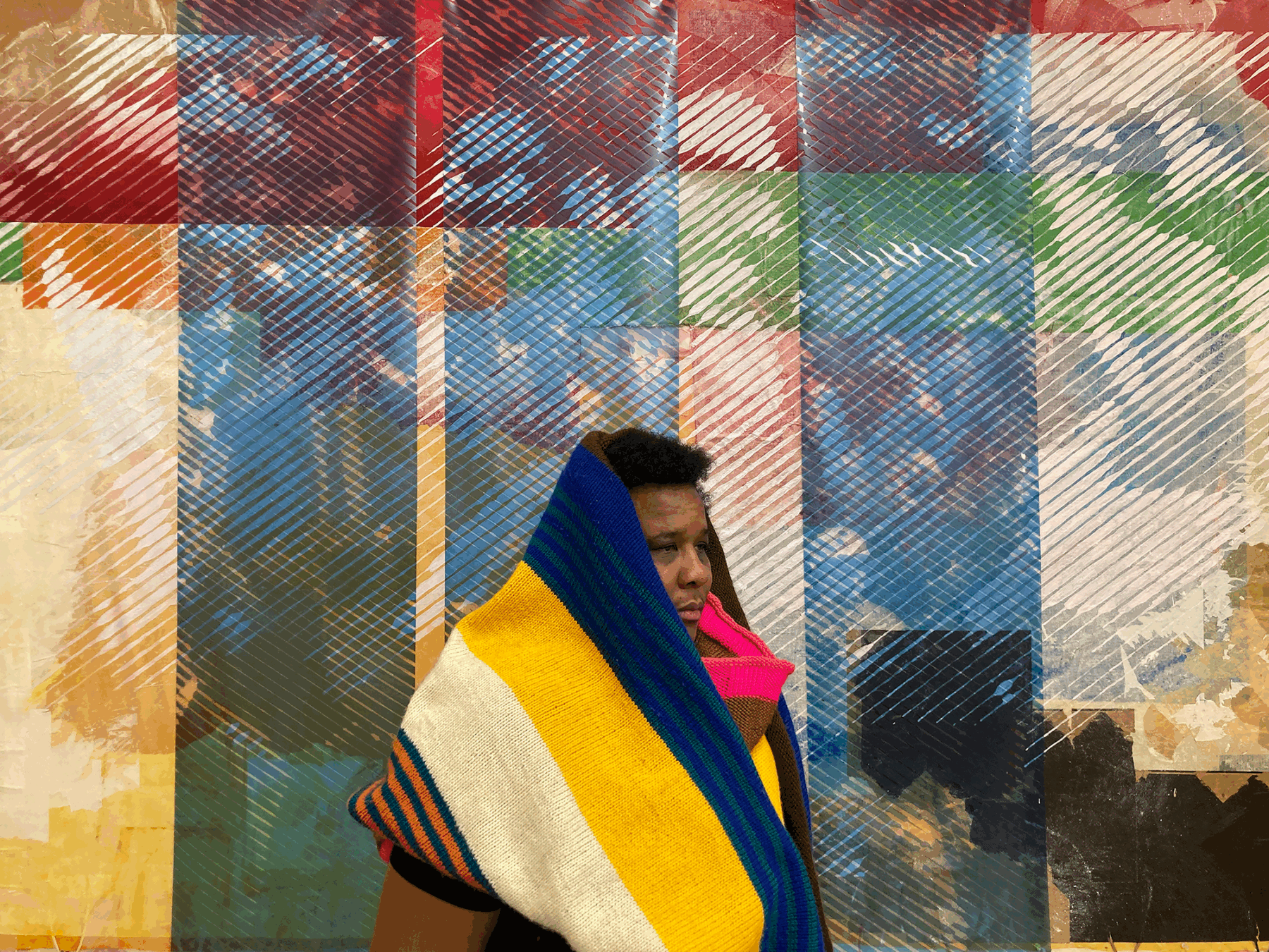 A woman with short black hair and a dark skin-tone stands with a blanket wrapped around her. She stands against a multicolor printed backdrop.