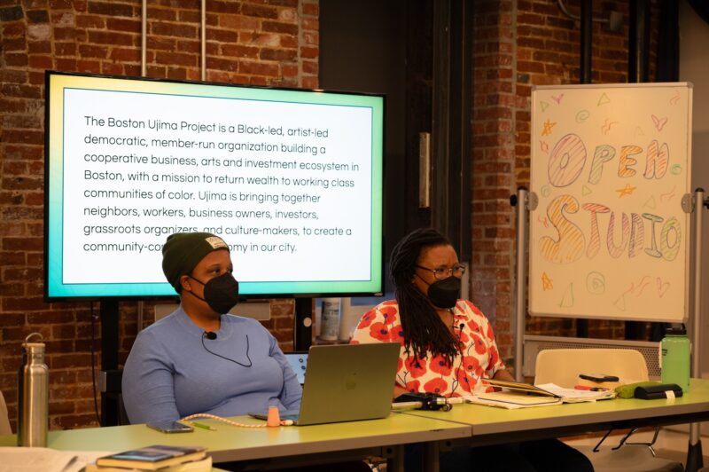 Two women with dark skin-tones and black face masks are seated at a table with a powerpoint projected behind them. The woman to the left wears an olive green beanie and a light blue long-sleeve shirt. At right, a woman with longer braids wears a red-and-white floral shirt.