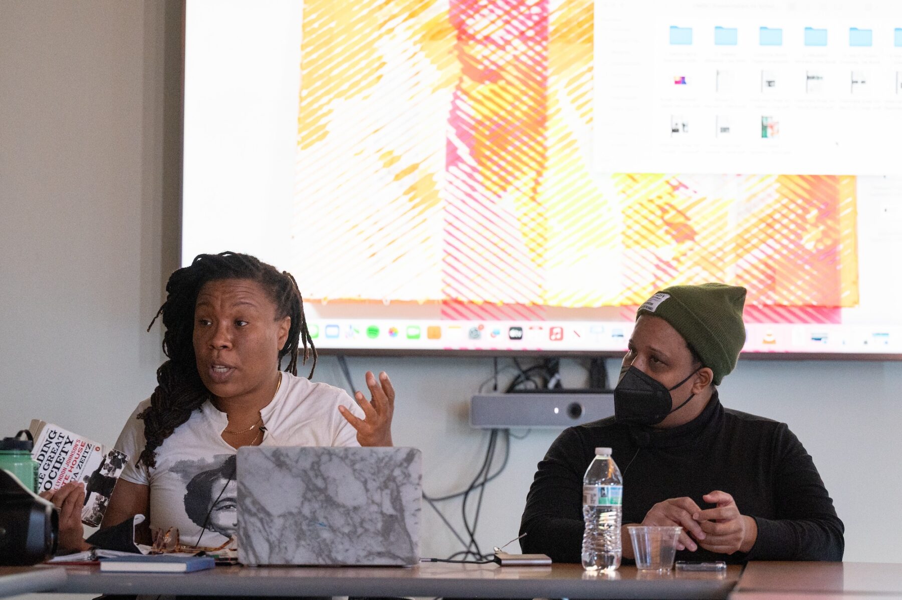 Two women with a dark skin-tone are seated at a wooden table. At left, a woman with longer braids and a white t-shirt sits in front of an open laptop and raises her hand in gesticulation. At right, a woman in an olive green beanie and a black button up shirt turns to face her companion.