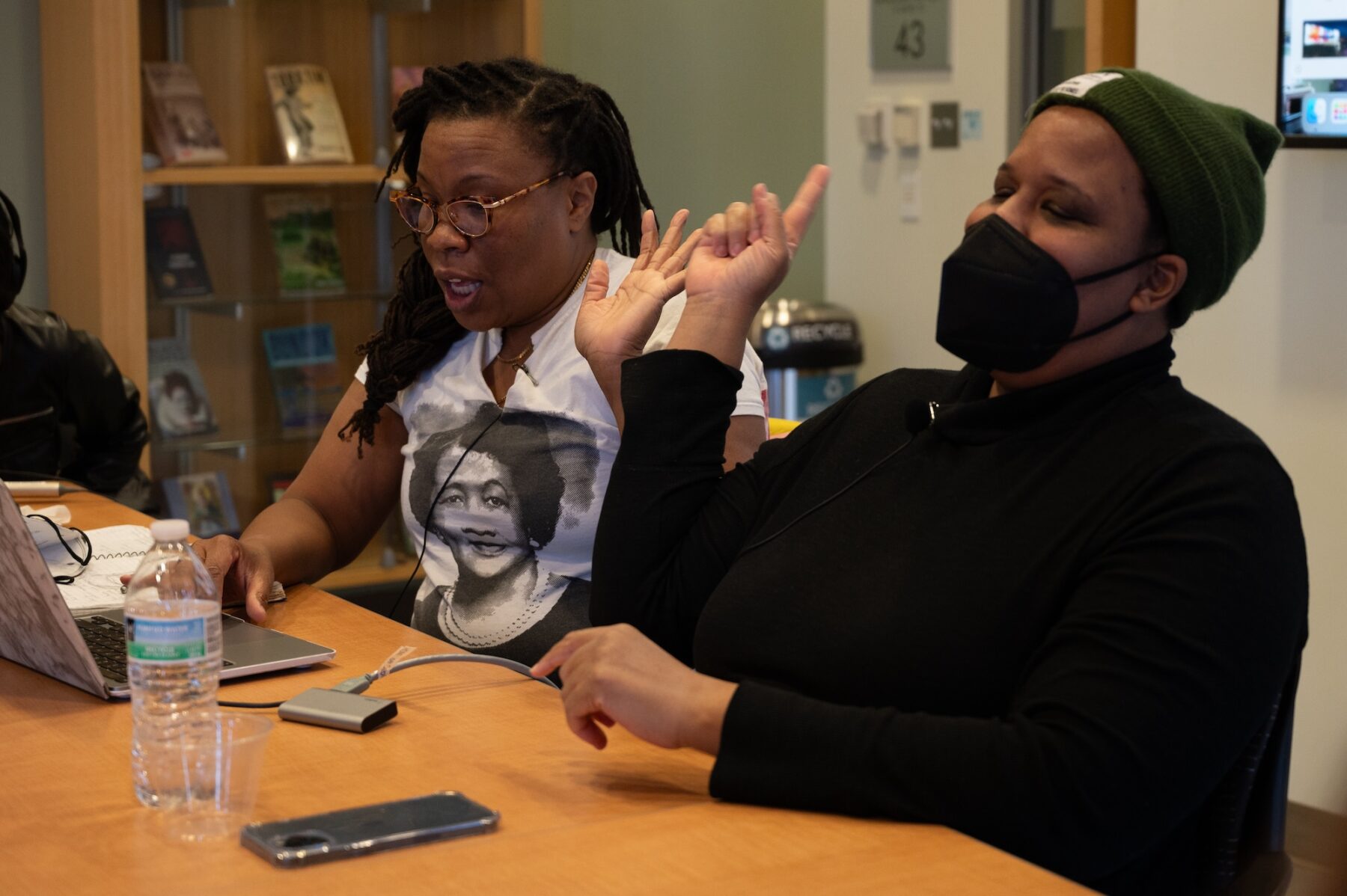 Two women with a dark skin-tone are seated at a wooden table. At left, a woman with longer braids and a white t-shirt sits in front of an open laptop. At right, a woman in an olive green beanie and a black button up shirt raises her right hand in a pointing motion.