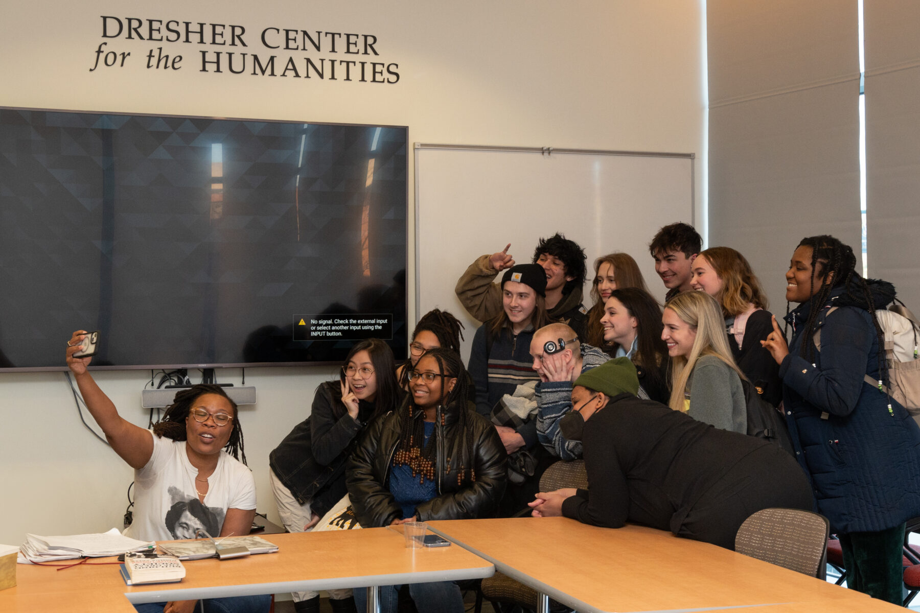 A woman with a dark skin-tone is seated before a larger group, holding up her cell phone to take a selfie. The woman wears tan glasses and a white t-shirt featuring a black-and-white photograph. The group behind her poses and smiles in anticipation of the photograph.
