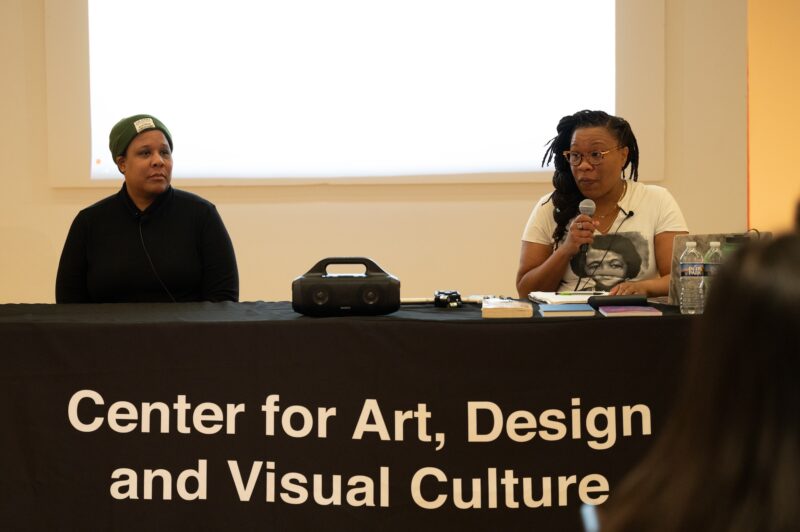 Two women sit behind a table with a black tablecloth reading "Center for Art, Design, and Visual Culture" in white text. At left is a woman with a dark skin-tone with an olive green beanie and a black button up shirt. To the right is a woman with a dark skin-tone with long braids and wearing a white t-shirt. She holds a microphone up to her mouth and is speaking.