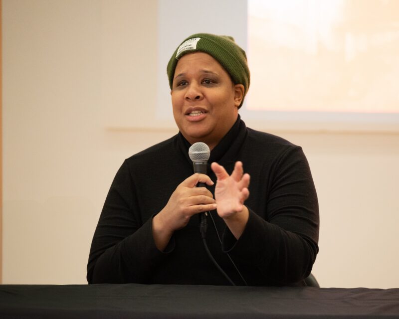 A woman with a dark skin-tone wears an olive green beanie and a black button up shirt. She holds a microphone up in her right hand and gesticulates with her left.