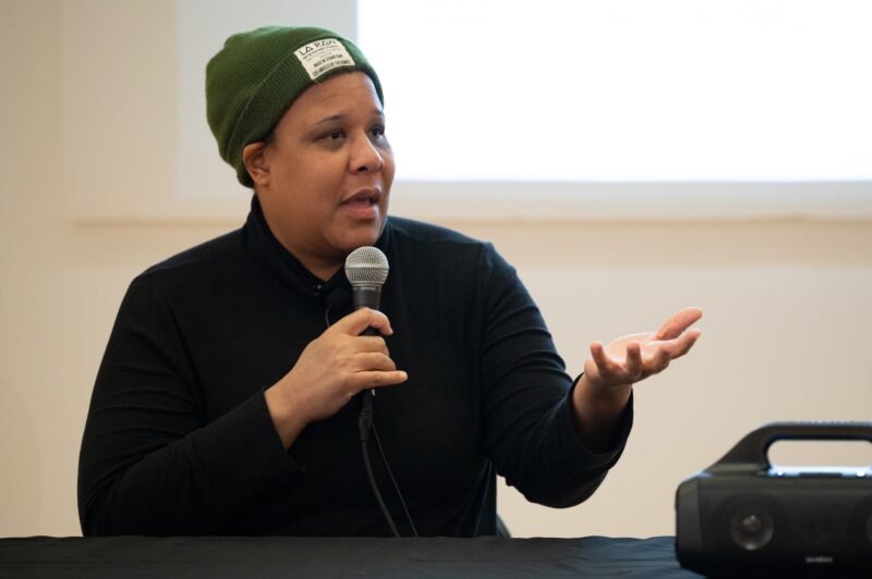 A woman with a dark skin-tone wears an olive green beanie and a black button up shirt. She holds a microphone up in her right hand and gesticulates with her left.