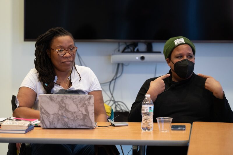 Two women with a dark skin-tone are seated at a wooden table. At left, a woman with longer braids and a white t-shirt sits in front of an open laptop. At right, a woman in an olive green beanie and a black button up shirt raises her hands to her masked face.