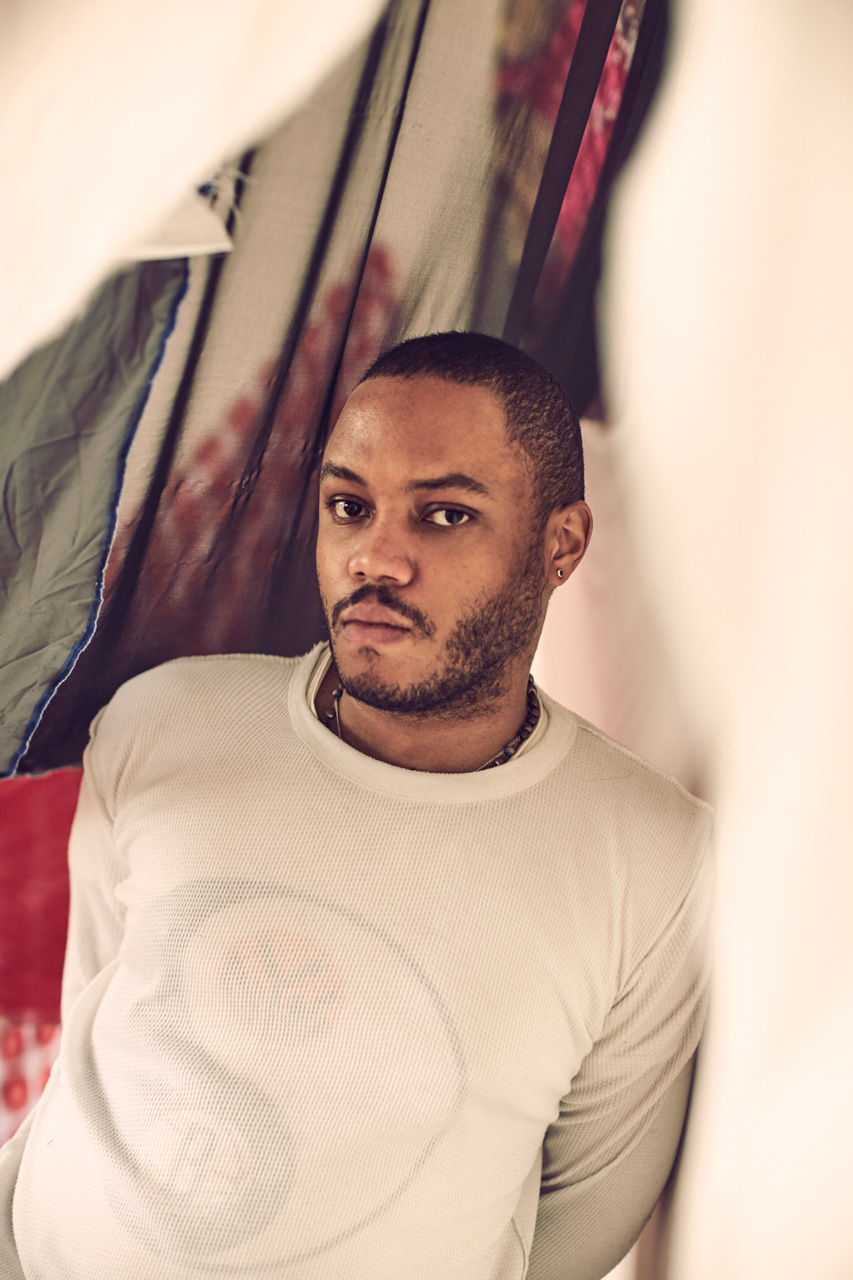 Eric N. Mack leans up against a white wall with a green, red, and cream colored textile hanging behind him.