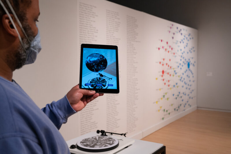 A individual with a dark skin-tone wearing a light-blue face mask holds up an iPad in front of a turntable.