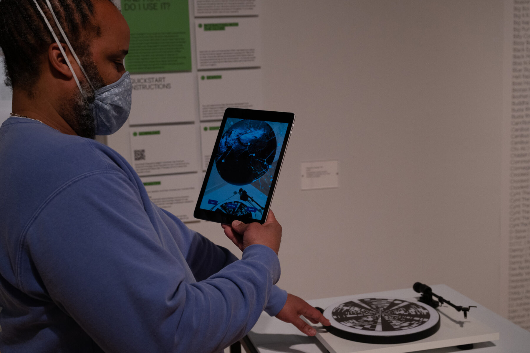 A individual with a dark skin-tone wearing a light-blue face mask holds up an iPad in front of a turntable.