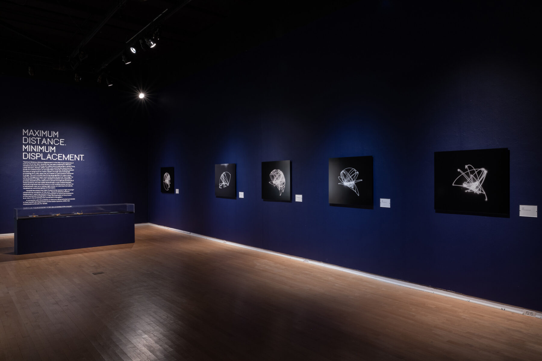 Installation view of a gallery space. The walls are dark navy, and five abstract works in black and white line the walls. On the back wall is didactic text with the title "Maximum Distance. Minimum Displacement." In front of this wall is a glass case with more work on display.