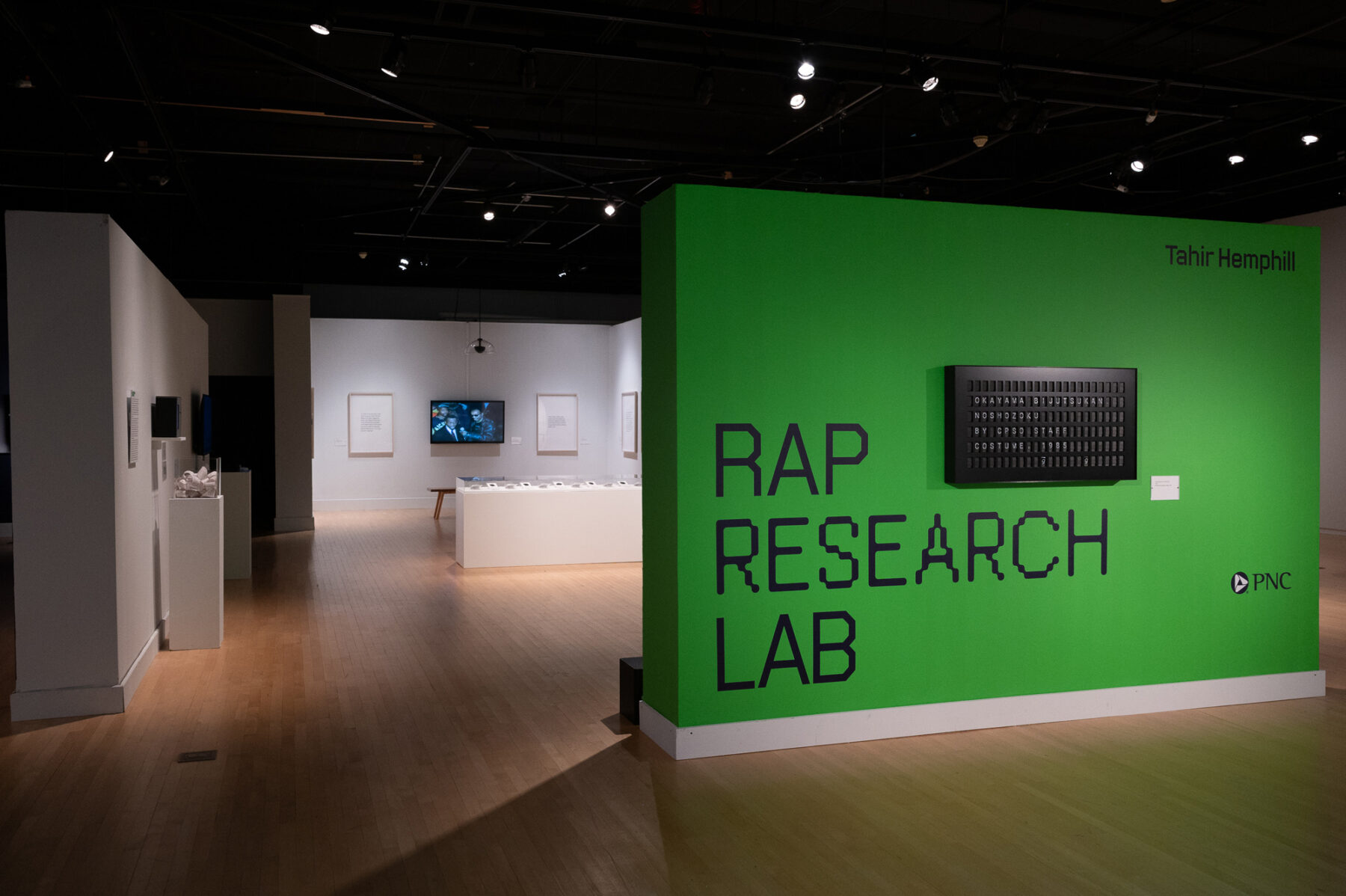 Installation view of Tahir Temphill: Rap Research Lab. A bright green wall takes up the foreground and reads "Tahir Temphill" and "RAP RESEARCH LAB." Behind this wall are various works of art installed in a white-walled gallery.