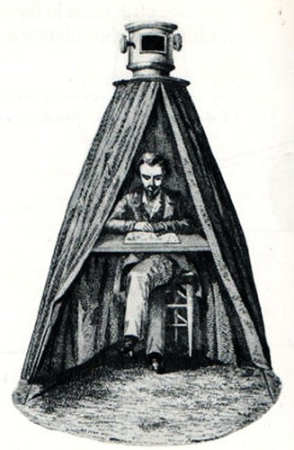 the camera obscura; a man sits at a desk in a triangle shaped tent with a device at the top that allows light in.