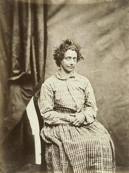 A sepia image shows a woman seated in front of a curtain. She has a long dress on that is striped and made of simple fabric like cotton or linen. Her hair is short and very curly.