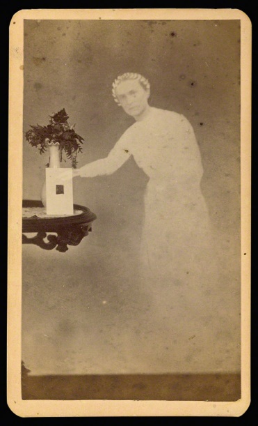 An old photograph shows a ghostly figure of a woman in a white dress that becomes more transparent as it goes down. She is next to an ornately carved dark wooden table with a glass inset that has flowers and a card.