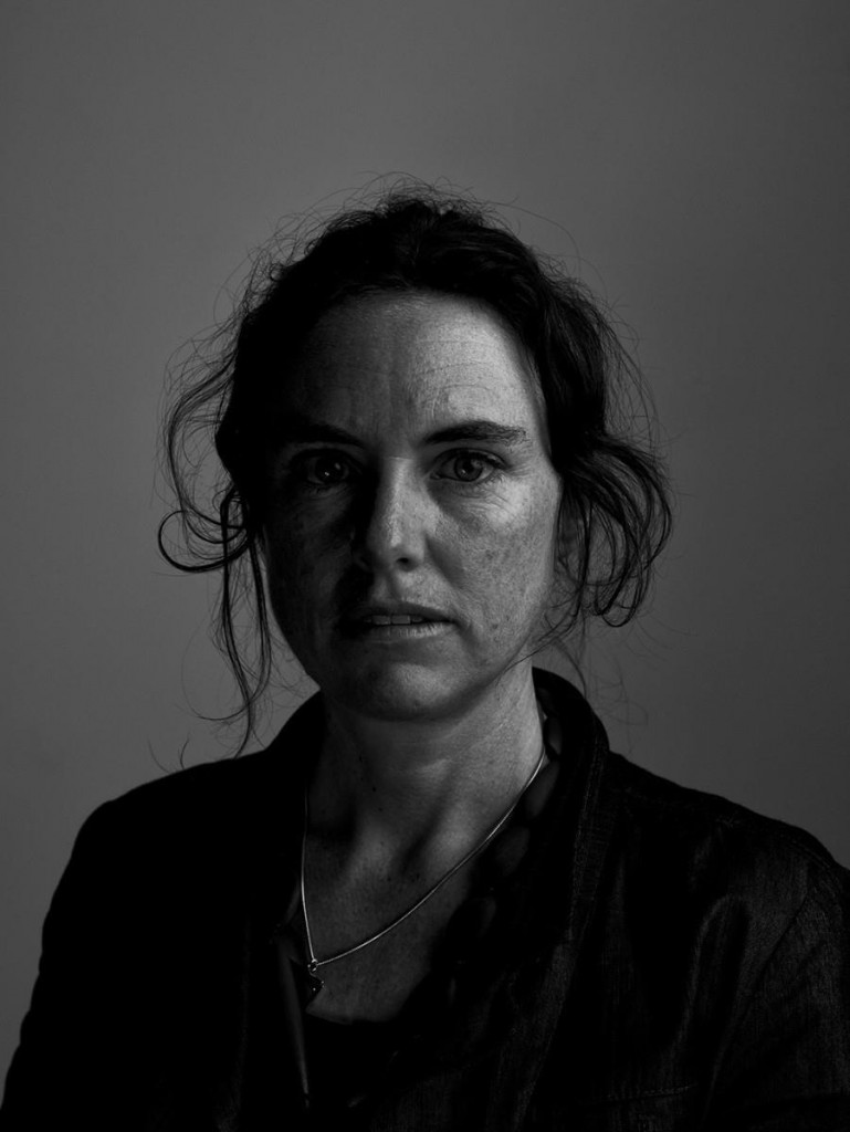 Black and white portrait of a woman with a serious expression, staring directly at the camera. Her hair is pulled back, with loose flyaways framing her face.