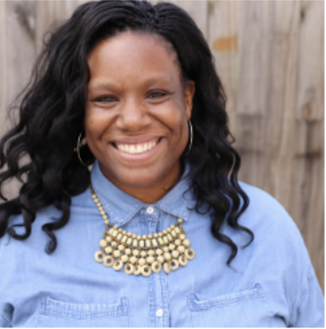 A portrait of a woman with a dark skin-tone with shoulder-length curly hair smiling at the camera. She wears an ornate gold necklace tucked into the collar of a light blue button-down shirt.