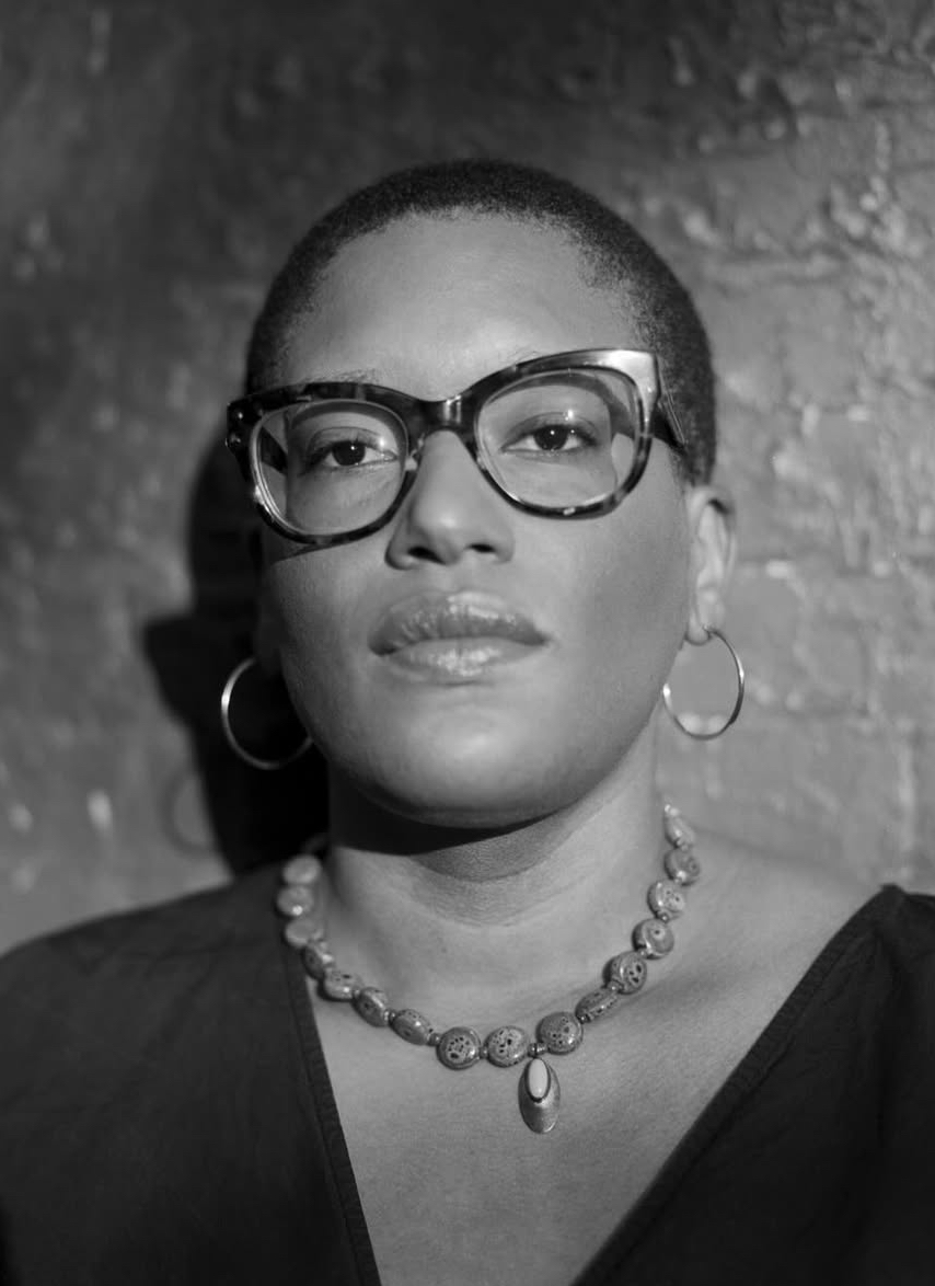A black-and-white portrait of a woman with a dark skin-tone. She has a shaved hairstyle and wears large cat-eye glasses, hoop earrings, and a larger metal necklace.