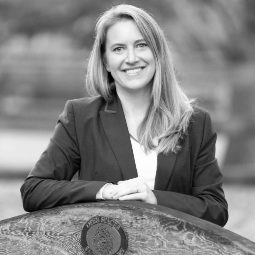 A black-and-white portrait of a woman with a light skin-tone smiling at the camera. She has shoulder-length blonde hair and is wearing a black blazer.