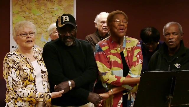 Above is a still image from the project introductory trailer. It shows a group of older Black and white men and women holding hands and singing together. There is a black music stand in front of them and a multi-colored map of the US on the dark red wall behind them.