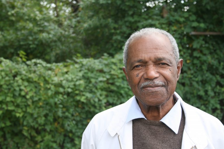 A portrait of a man with a dark skin-tone and a mustache. He wears a white suit jacket over a brown sweater vest and white collared shirt. The man is softly smiling and standing outside in front of greenery.