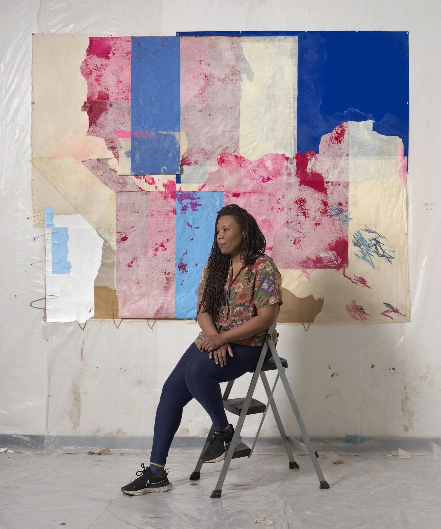 A portrait of a woman with a dark skin-tone seated on a step ladder. She is seated in front of a mixed media work of art mounted on a wall.
