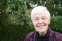 A portrait of a senior woman with a light skin-tone. She has short white hair and wears thin glasses, a black turtleneck, and a purple sweater. The woman is smiling and standing outside in front of greenery.
