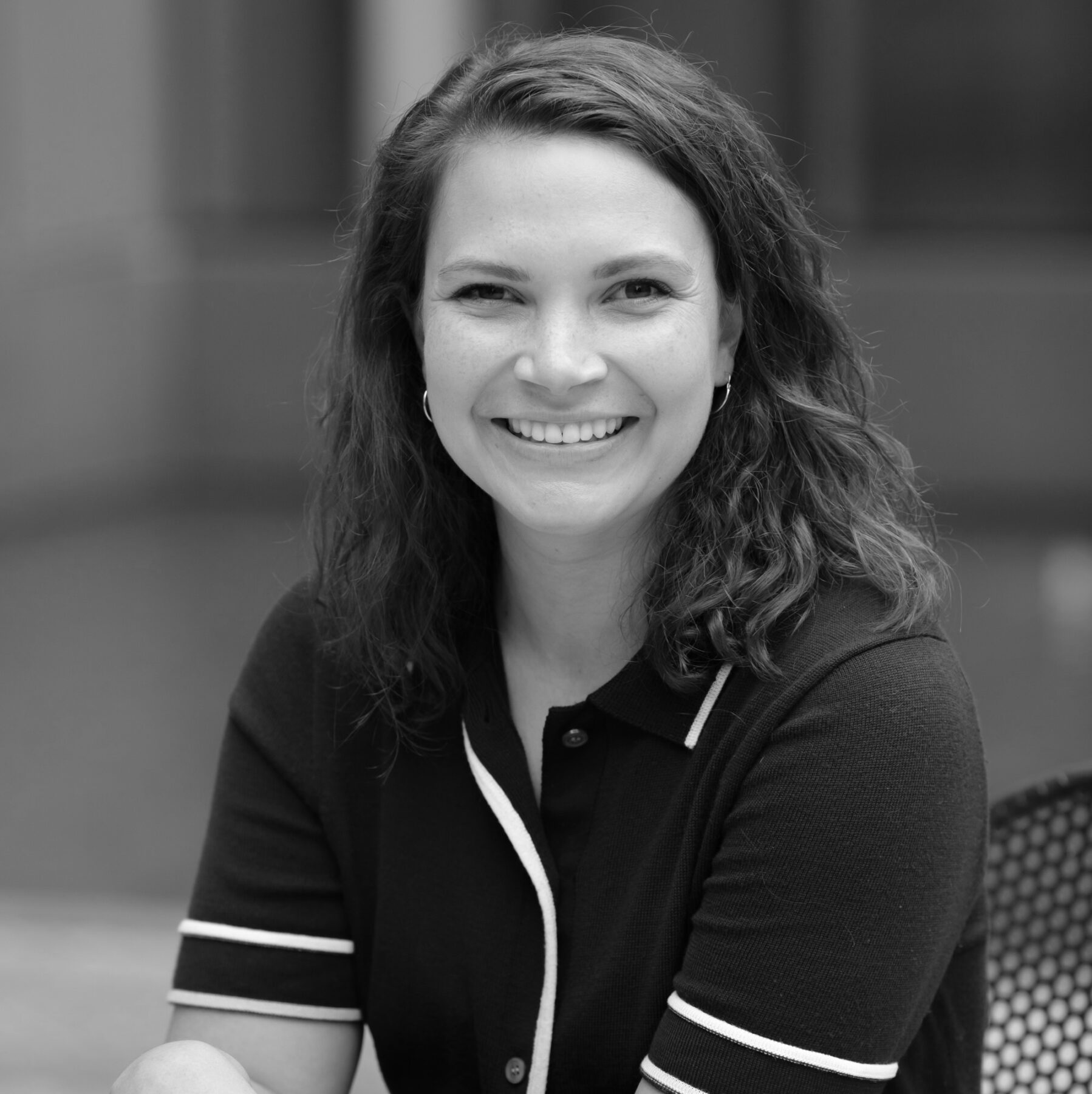 A black-and-white portrait of a woman with a light skin-tone who smiles at the camera. She has medium-length dark-colored wavy hair and wears a dark shirt with white piping.