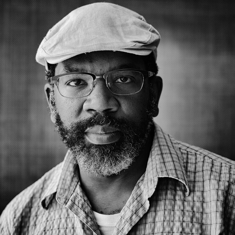 Black-and-white portrait of a man with a dark skin-tone and a beard. He is wearing a pale-colored newsboy has, wire-rimmed classes, and a patterned button up shirt.
