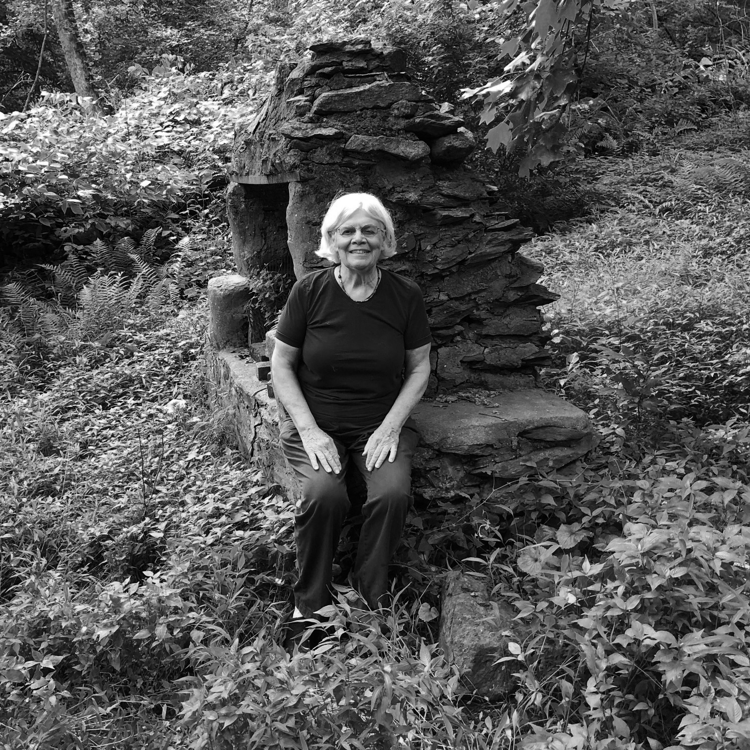 A black-and-white portrait of an elderly woman with a light skin-tone seated and looking at the camera. She has a grey-haired bob and wears a black outfit. She rests her hands on her knees. She is surrounded by a wild garden.