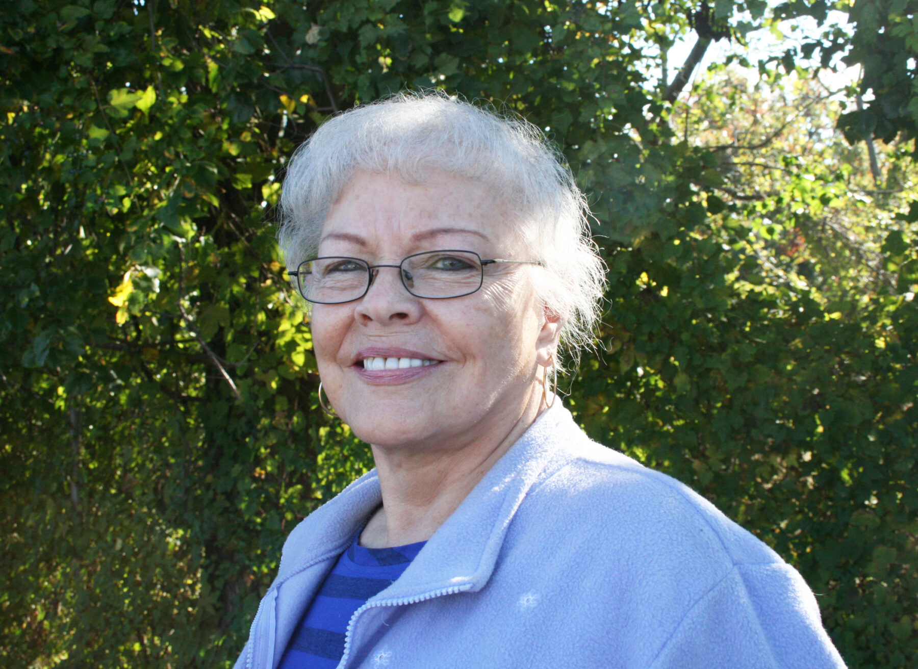 A portrait of a senior woman with a light skin-tone. She has short white hair and wears thin glasses and a blue cardigan over a striped blue shirt. The woman is smiling and standing outside in front of greenery.