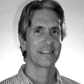 A black-and-white portrait of a man with a with a light skin-tone with dark colored hair. He smiles for the camera and wears a gingham-print shirt.