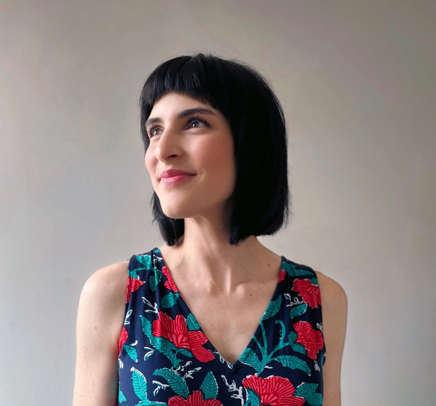 A portrait of a woman with a light skin-tone and a brown bob. She wears a blue and red floral blouse and looks over her right shoulder away from the camera.