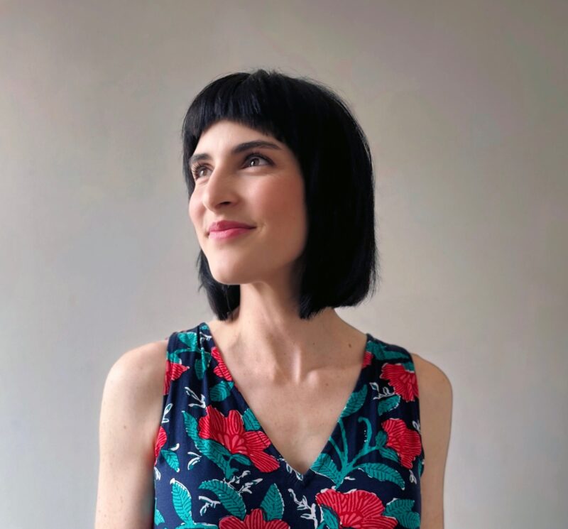 A portrait of a woman with a light skin-tone and a brown bob. She wears a blue and red floral blouse and looks over her right shoulder away from the camera.