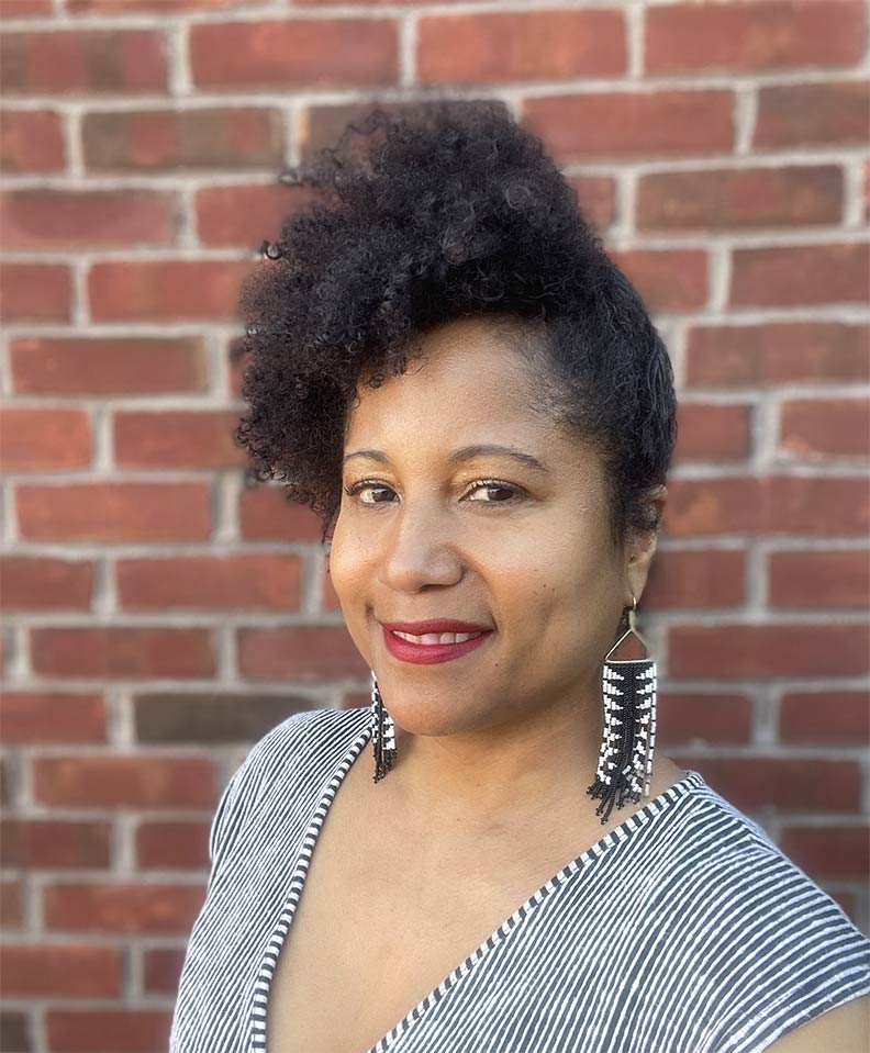 Dell Marie Hamilton's headshot; she has light to medium skin tone and wears earrings and a v-neck shirt in front of a brick wall.