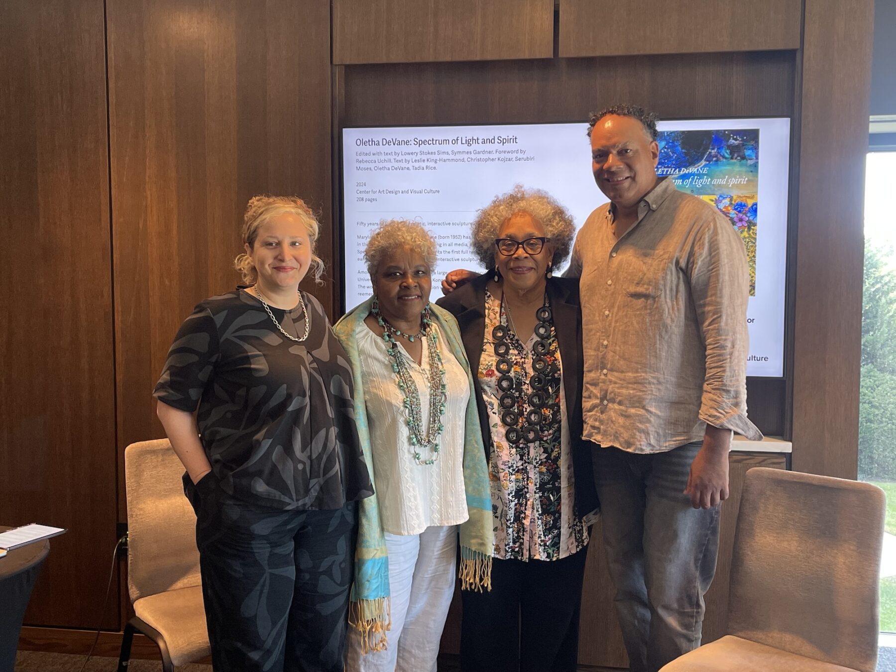 Four individuals (three woman and a man to the far right) stand facing the camera, smiling with their arms around each other. Behind them is a screen featuring a powerpoint slide.