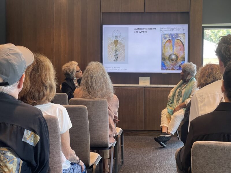 A seated audience in grey chairs face the front of a room. Two older women with dark skin-tones engage in discussion with a powerpoint slide projected behind them.