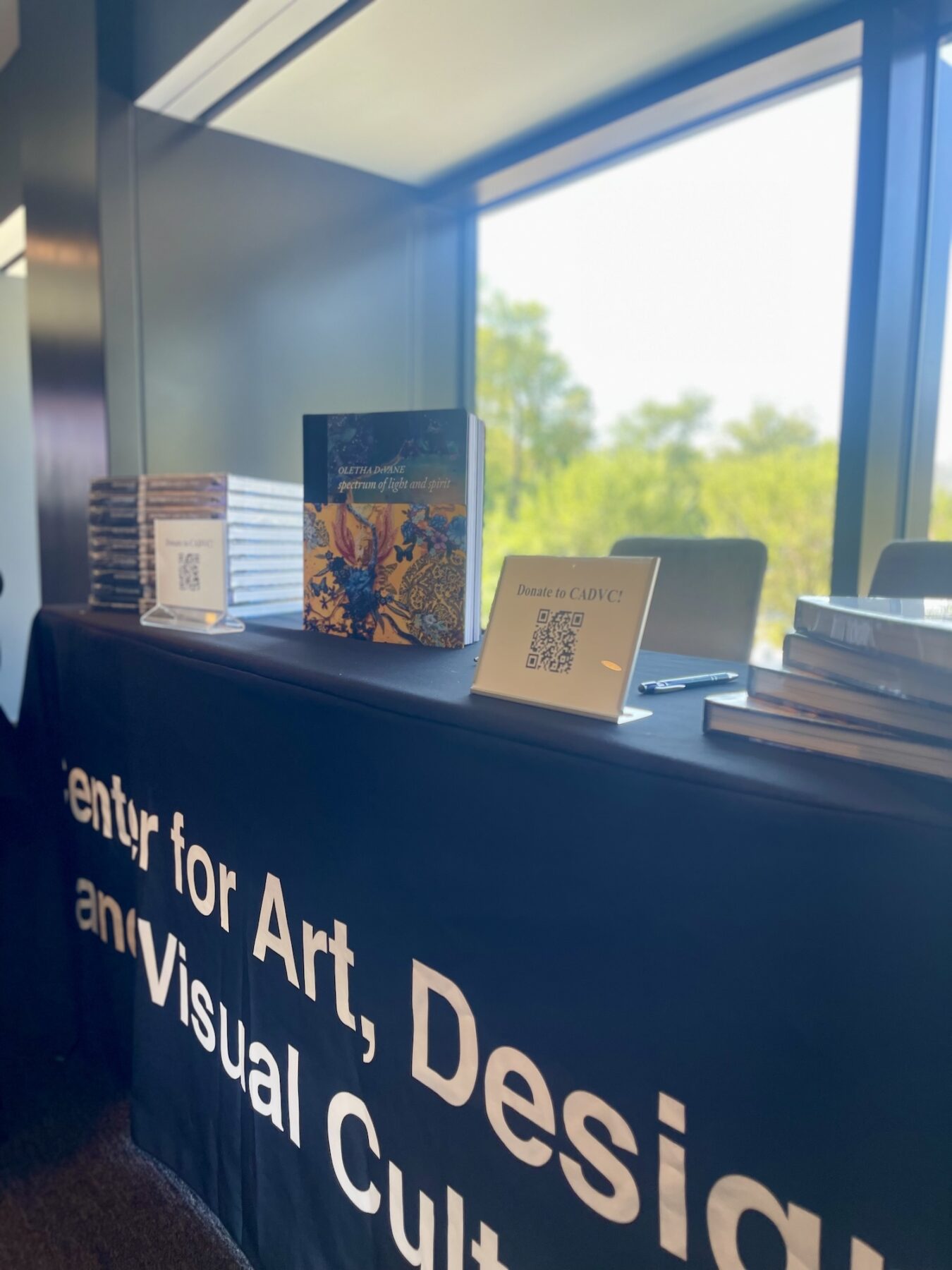 A table is decorated with a black table cloth and stacks of books with one faced out for display. The skirt of the table cloth is decorated with white lettering: "Center for Art, Design, and Visual Culture."