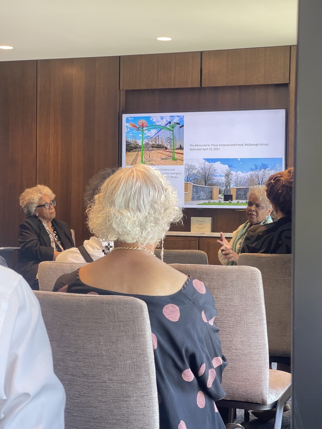 A seated audience in grey chairs face the front of a room. Two older women with dark skin-tones engage in discussion with a powerpoint slide projected behind them.