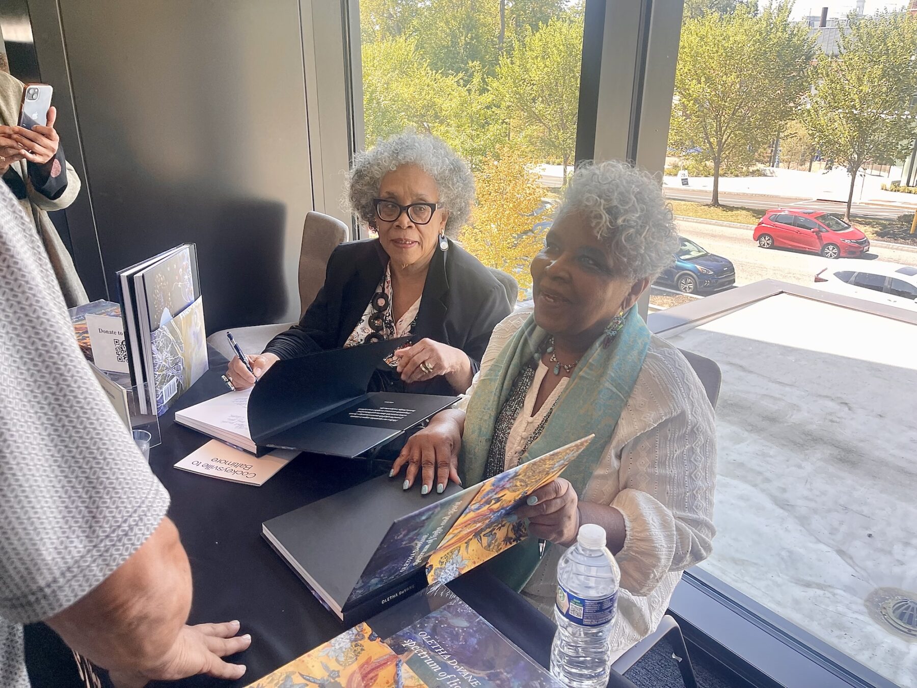 Two older women with dark skin-tones are seated at a table for a book signing. The woman closest to the camera wears a white blouse with a teal scarf. She looks up and smiles at someone standing before her on the other side of the table.