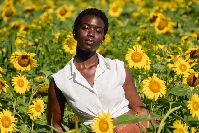 M'Balou Camara's headshot; she has a darker skin tone and wears earrings and a collared shirt. She is outside on a sunny day in a field of sunflowers.