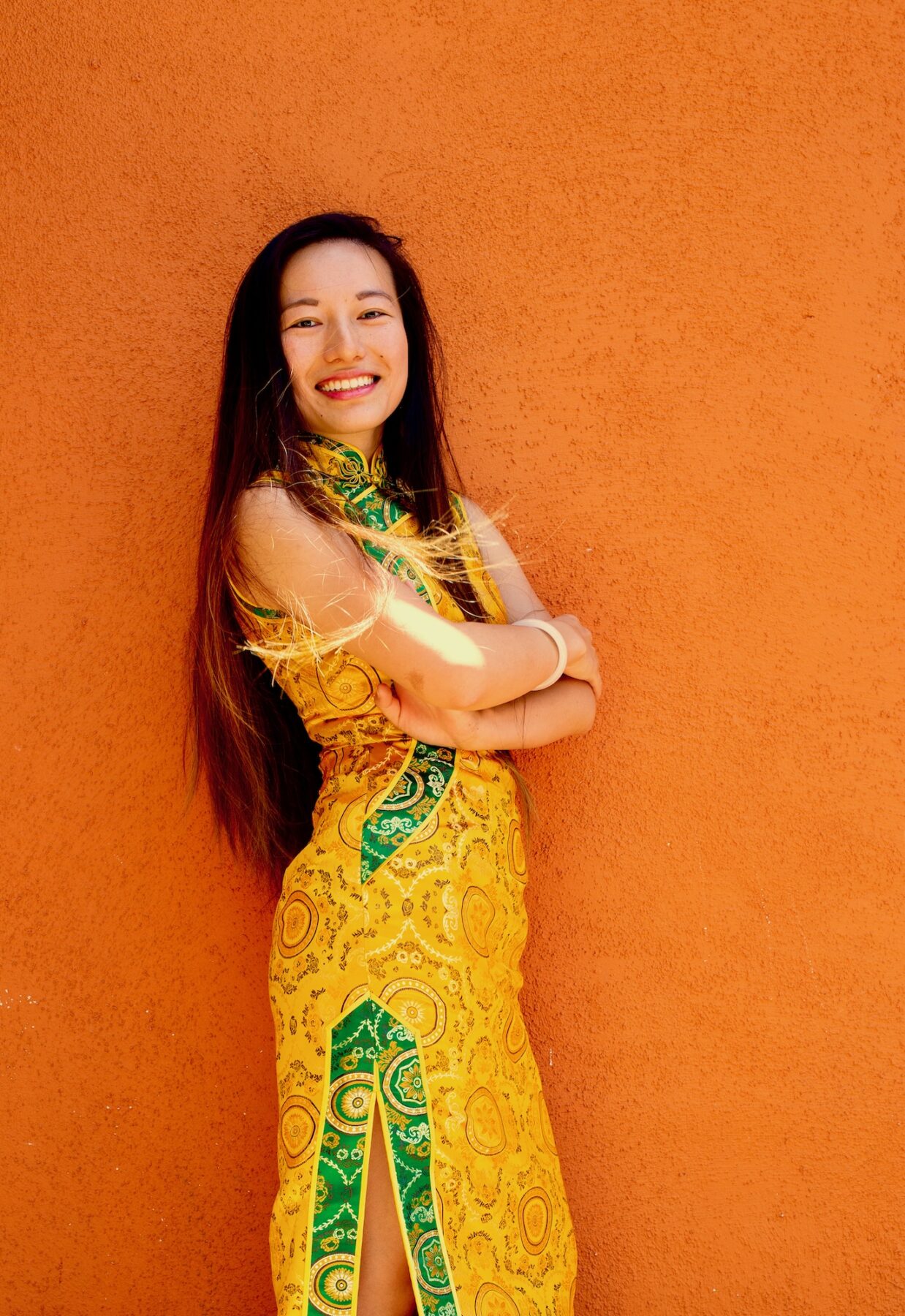 A photo of an Asian woman with a medium skin-tone standing against an orange wall with her arms crossed. She has long brown hair and wears an ornate yellow and green dress.