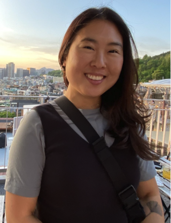 Photo of an Asian woman with a medium skin-tone standing outside. She wears a grey shirt, a black vest, and a black crossbody bag. She has medium-length hair and smiles at the camera.