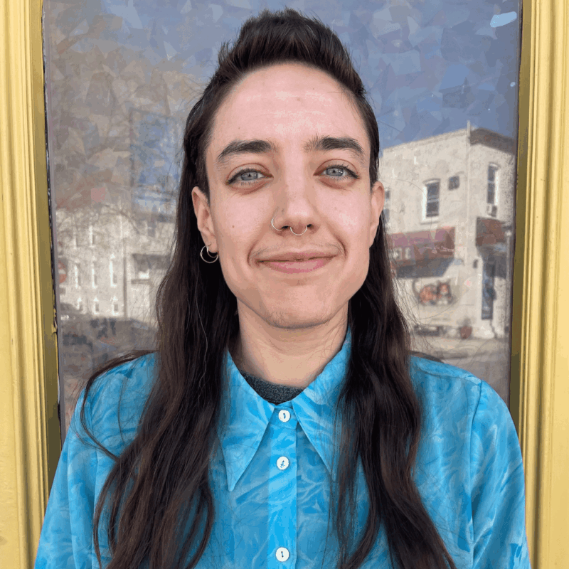 A portrait of an individual with a light skin-tone and long brown hair. They grin for the camera and wear a teal button up. They stand in front of a work of art in a gold frame.
