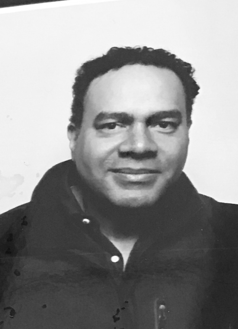 A black-and-white portrait of a man which a medium skin-tone grinning for the camera. He wears a black jacket and his black curly hair is cut short.