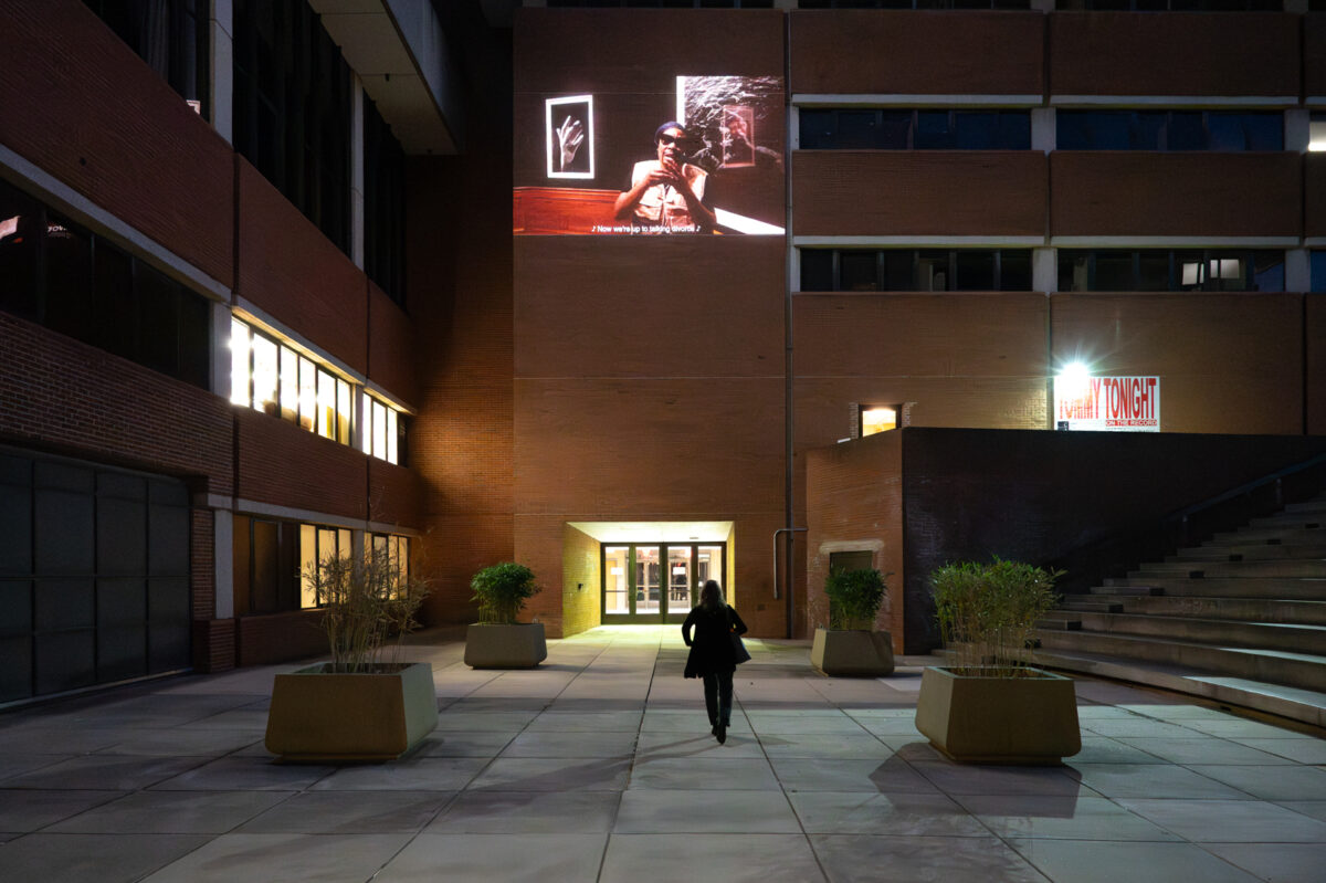 A silhouette of an audience member standing in the CADVC amphitheater at night, looking upward at a projected video showing Tommy Tonight, a drag performer wearing sunglasses and singing to the camera, with a gallery in the background.