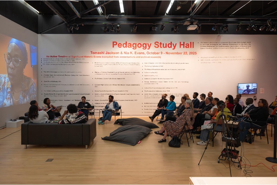 Large gallery space with a video projection of Tomashi Jackson on the far left wall, a circle sofa seating and an audience of 30 people facing those seated on the sofas. The back wall has a mural of a black text with bullet points illustrating a timeline of significant historic events compiled from conversation and archival research. The title, "Pedagogy Study Hall Tomashi Jackson & Nia K. Evans, October 9 - November 22, 2025" appears at the top in a large red sans serif font. A TV monitor below glows with a person depicted and a camera on tripod is set up in the foreground.